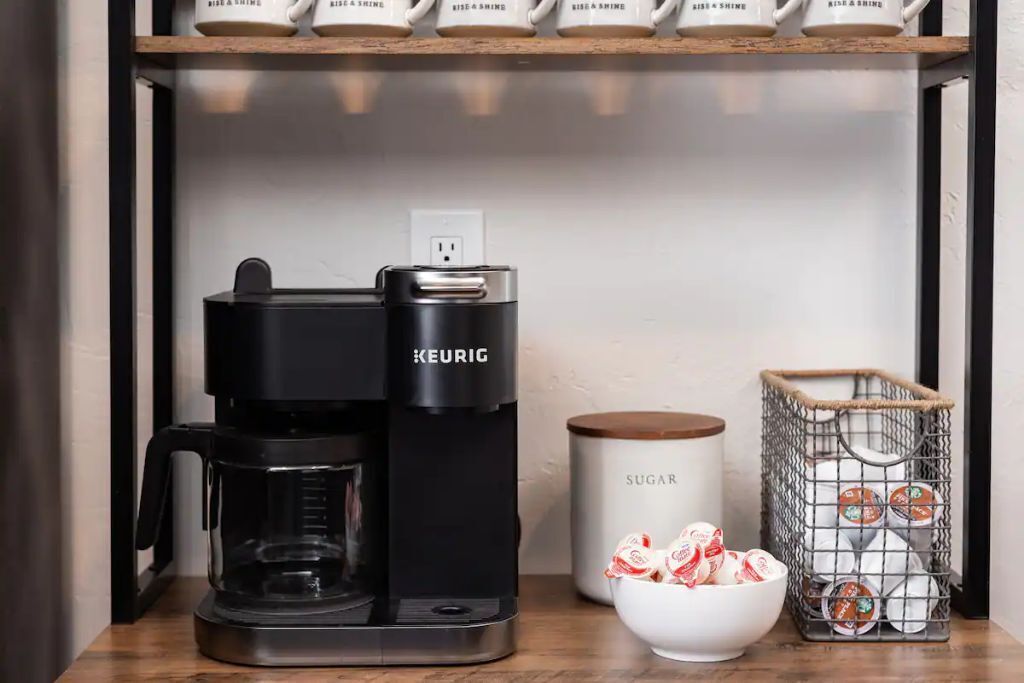 A Keurig coffee maker on a wooden shelf with a white mug labeled "RISE & SHINE" and a white sugar container.