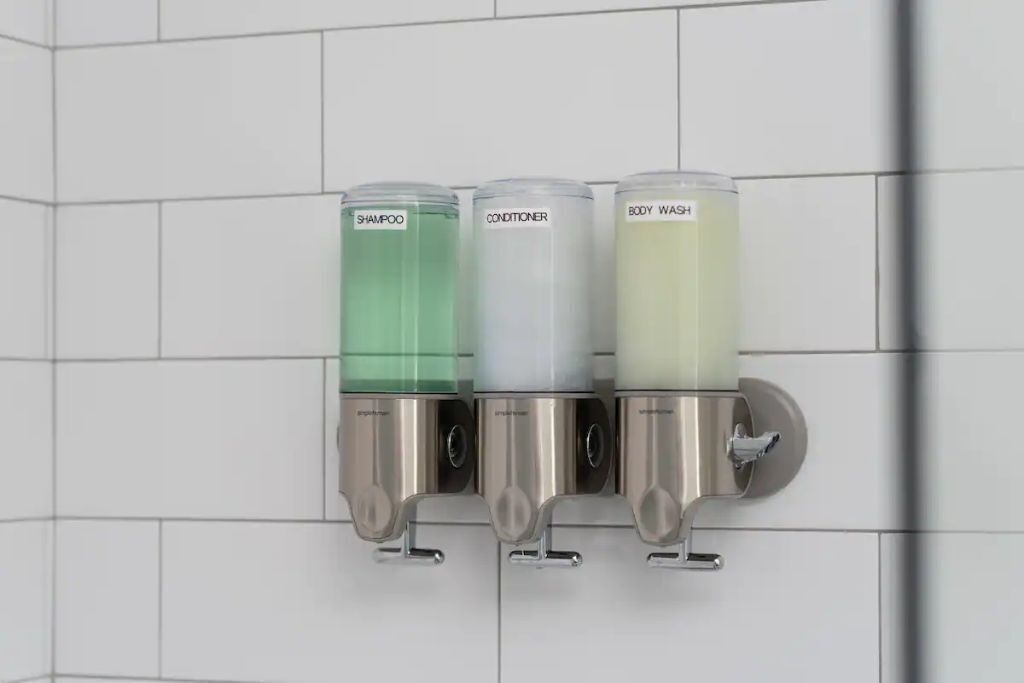 Dispensador mural con tres compartimentos etiquetados para champú, acondicionador y gel de baño.