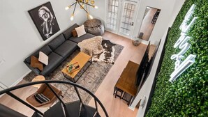 Uma sala de estar moderna com um sofá cinzento, uma mesa de centro de madeira e uma cadeira preta. Há uma obra de arte emoldurada a preto e branco na parede, um candeeiro único e uma parede verde com um jardim vertical.