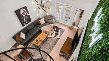 Un salon moderne avec un canapé gris, une table basse en bois et un fauteuil noir. Un tableau encadré en noir et blanc orne le mur, ainsi qu’un lustre original et un mur vert agrémenté d’un jardin vertical.