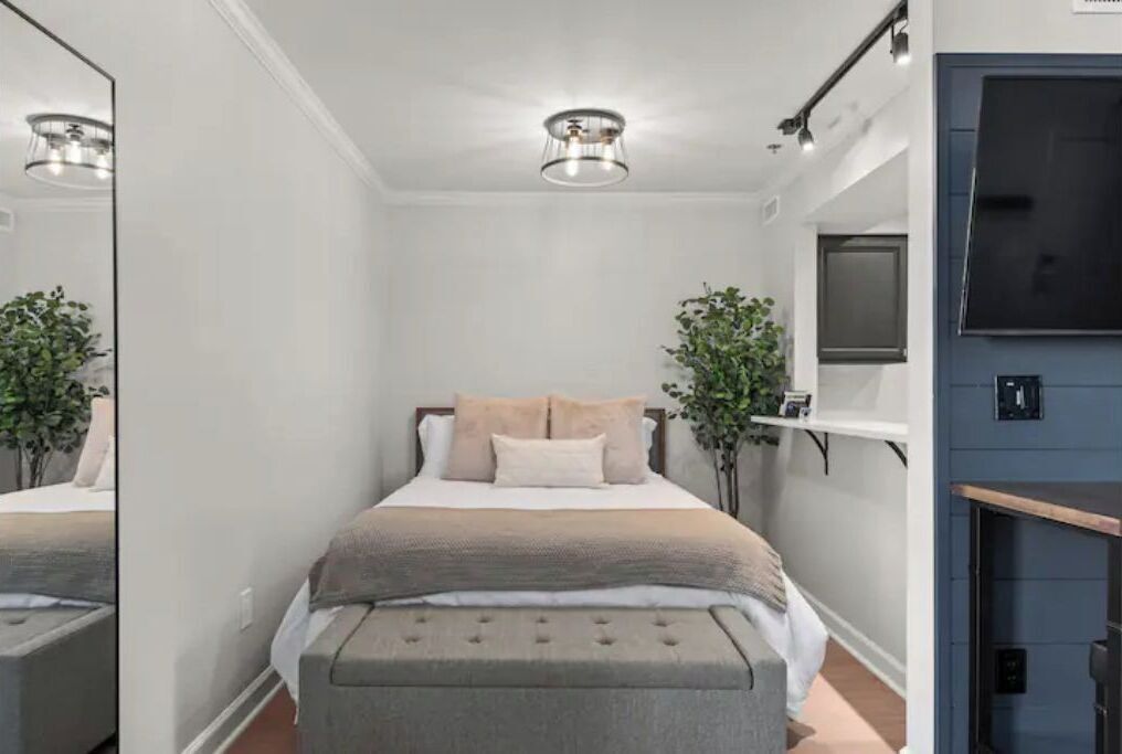 A narrow hotel room with a bed, a bench, a TV, and a mirror.