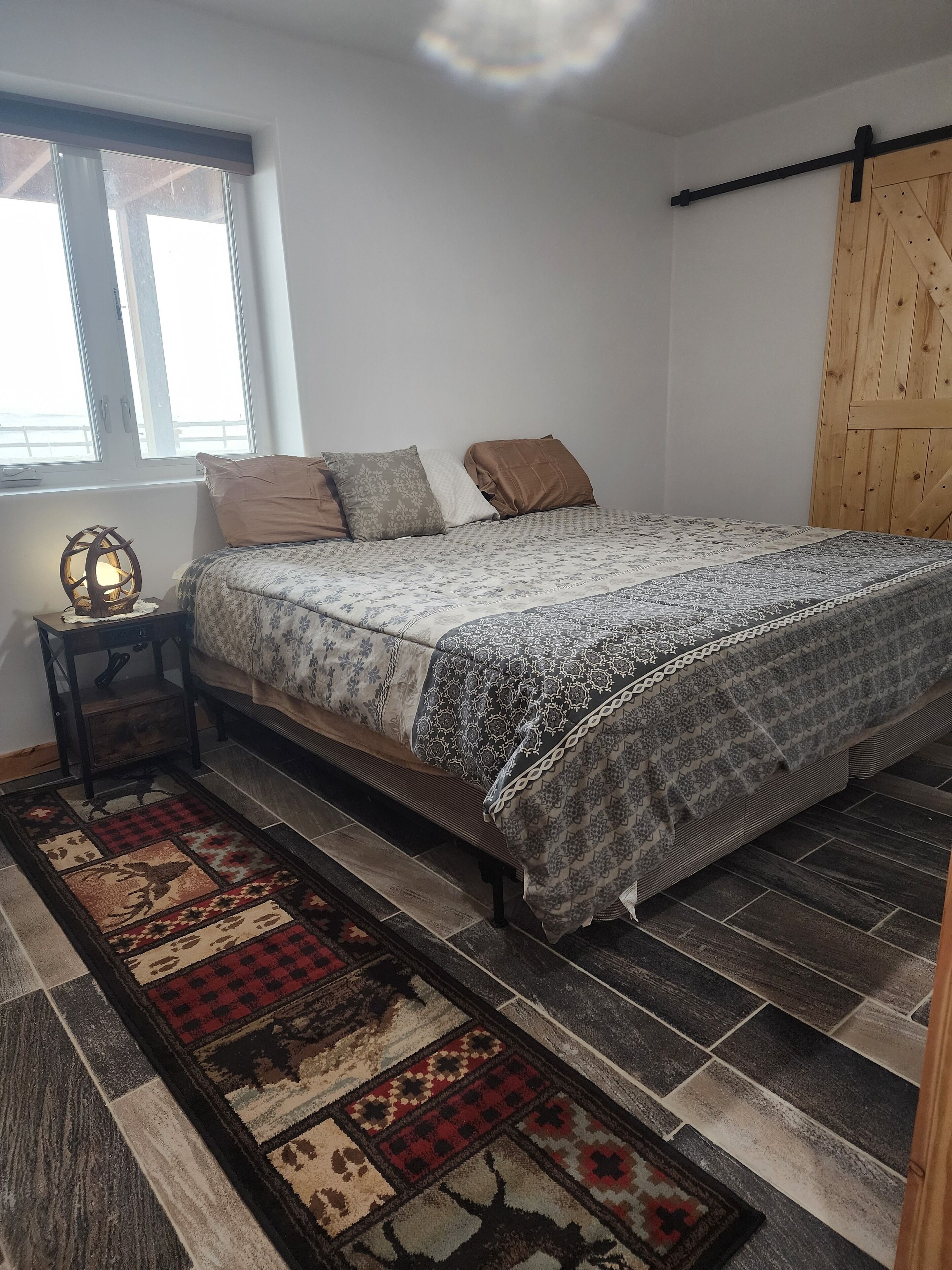 A bedroom with a bed, a nightstand, a lamp, a window, and a wooden door.