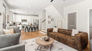 Una sala moderna con un sofá gris, un sofá de cuero marrón y una mesa de centro. Se ve una cocina al fondo con armarios blancos y un comedor con sillas.