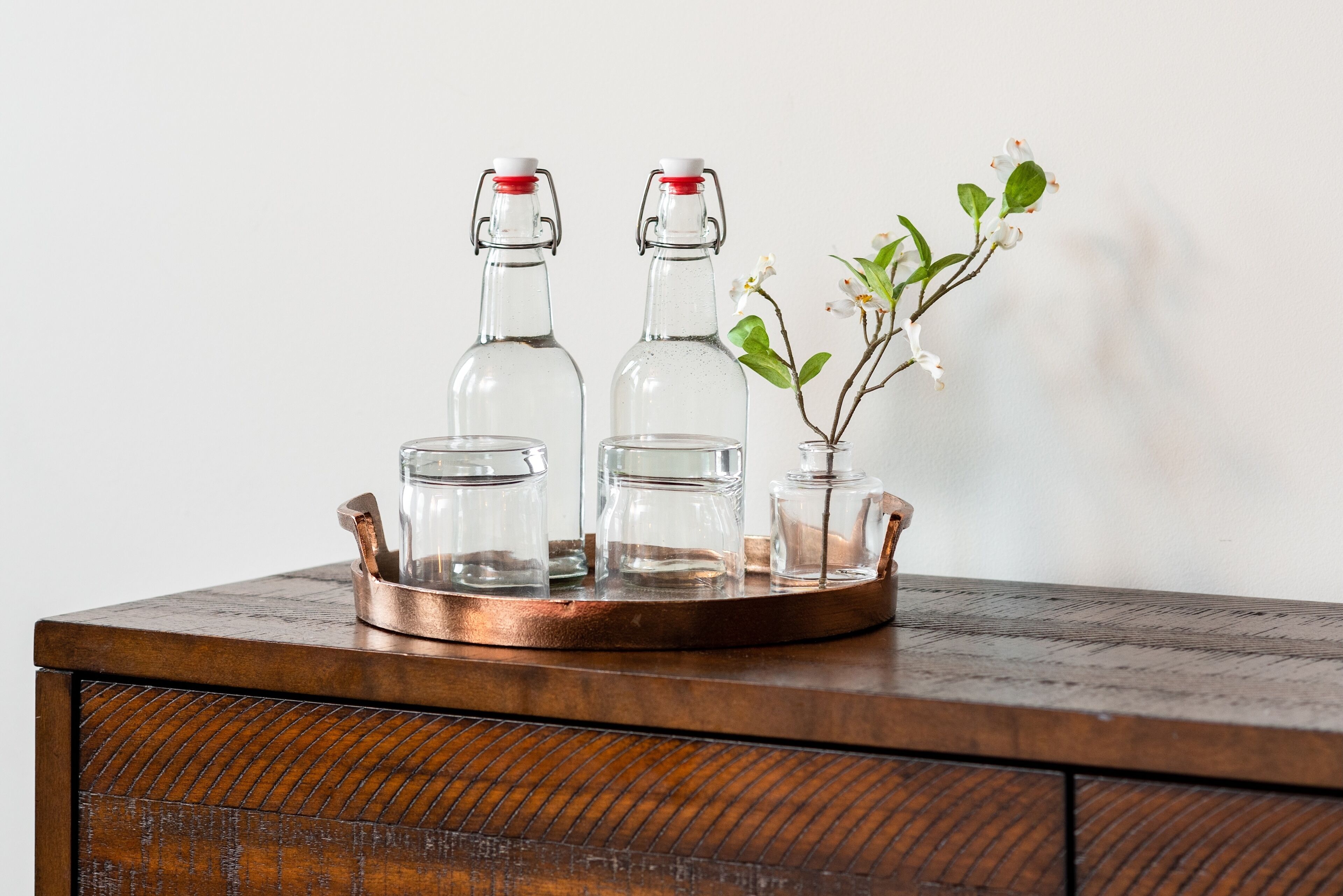 A wooden tray with three glass bottles and a small vase with flowers on a wooden surface.