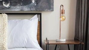 Una cama bien hecha con ropa blanca, un escritorio moderno con una lámpara y un cuadro en la pared.