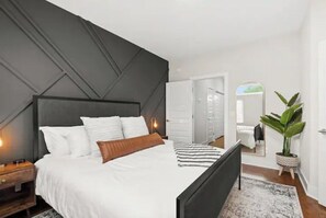 Quarto moderno com cama grande, cabeceira escura, mesa de cabeceira, espelho e planta em vaso.