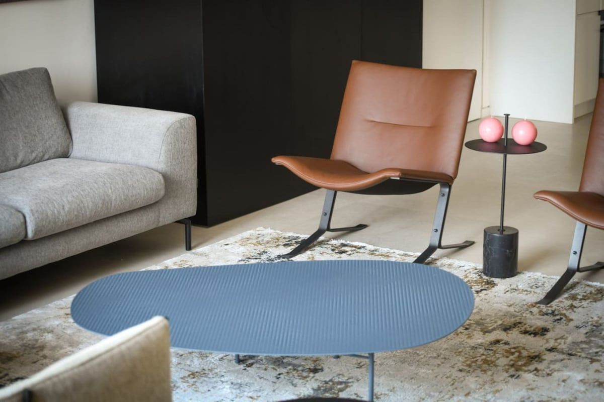A modern living room with a sofa, a brown leather chair, a small round table with two pink ornaments, and a textured rug.