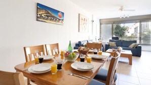 A dining area with a table set for two, a bowl of fruit, and glasses of juice. There is a sofa and a window in the background. - Bright 3BR Stay • Relaxing Comfort (Tel Aviv-Yafo)