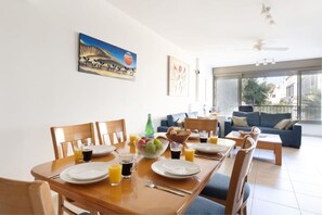 Un'area pranzo con un tavolo apparecchiato per due persone, una ciotola di frutta e bicchieri di succo. Si vede un divano e una finestra sullo sfondo.