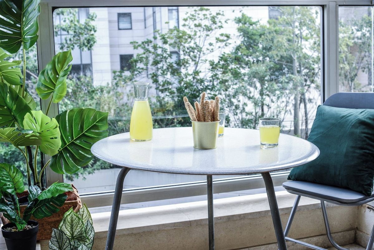 A round table with a glass pitcher and two glasses of yellow liquid, a container with sticks, and a green cushion on a balcony with a view of buildings and trees.