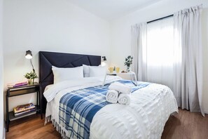 Un lit bien fait, recouvert d’une couverture à carreaux bleus et blancs, avec une tête de lit sombre et des oreillers blancs. On aperçoit une table de chevet avec une lampe, une petite plante et un plateau contenant divers objets. Une fenêtre avec des rideaux est visible à l’arrière-plan.