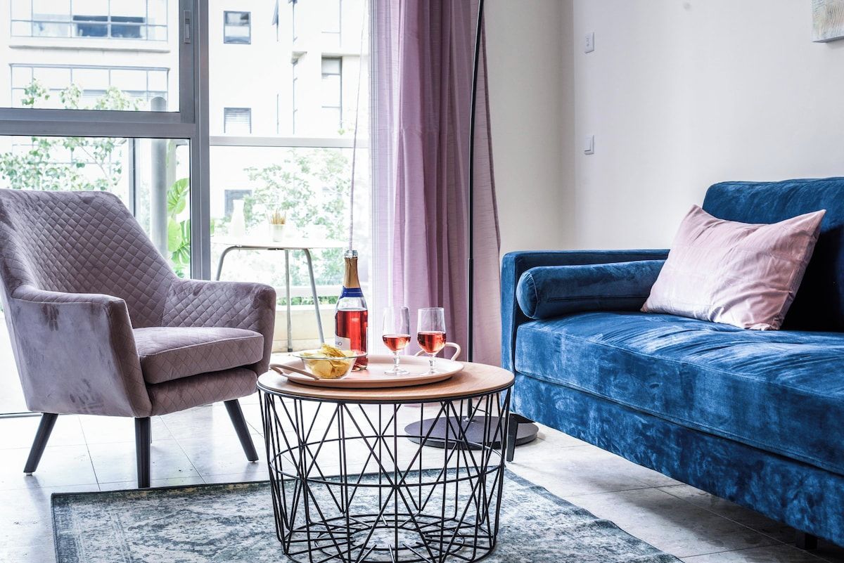Una sala de estar moderna con un sofá azul, un sillón cómodo, una mesa de centro redonda con una bandeja de aperitivos y bebidas, y vistas a edificios a través de amplios ventanales.