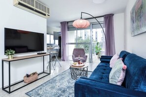 Un salon moderne avec un téléviseur à écran plat, un canapé bleu, une table basse ronde et une grande fenêtre avec des rideaux.