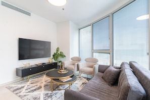 A modern living room with a flat-screen TV, a round coffee table, a sofa, and a large window with blinds.
