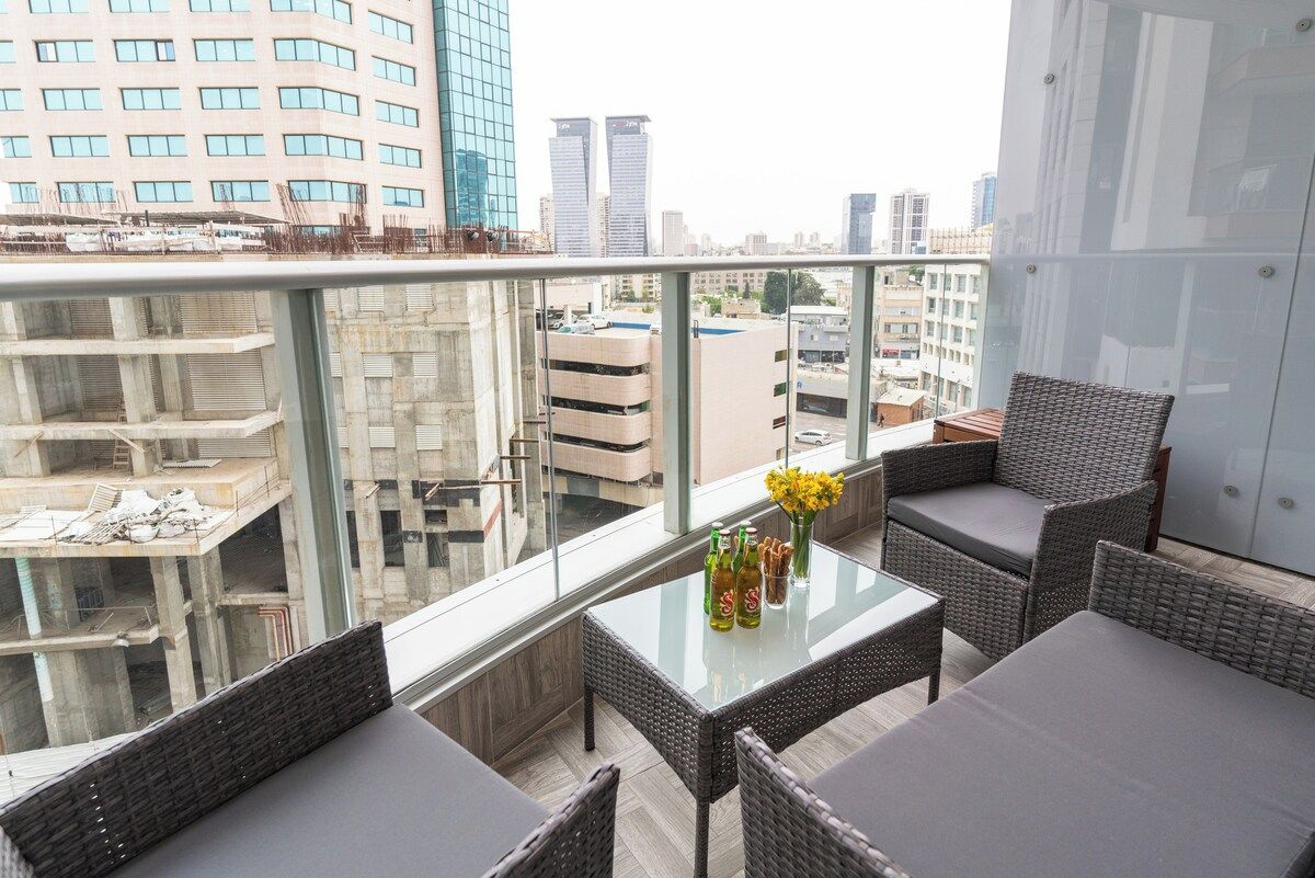 A balcony with wicker furniture, a glass table, and a view of city buildings.