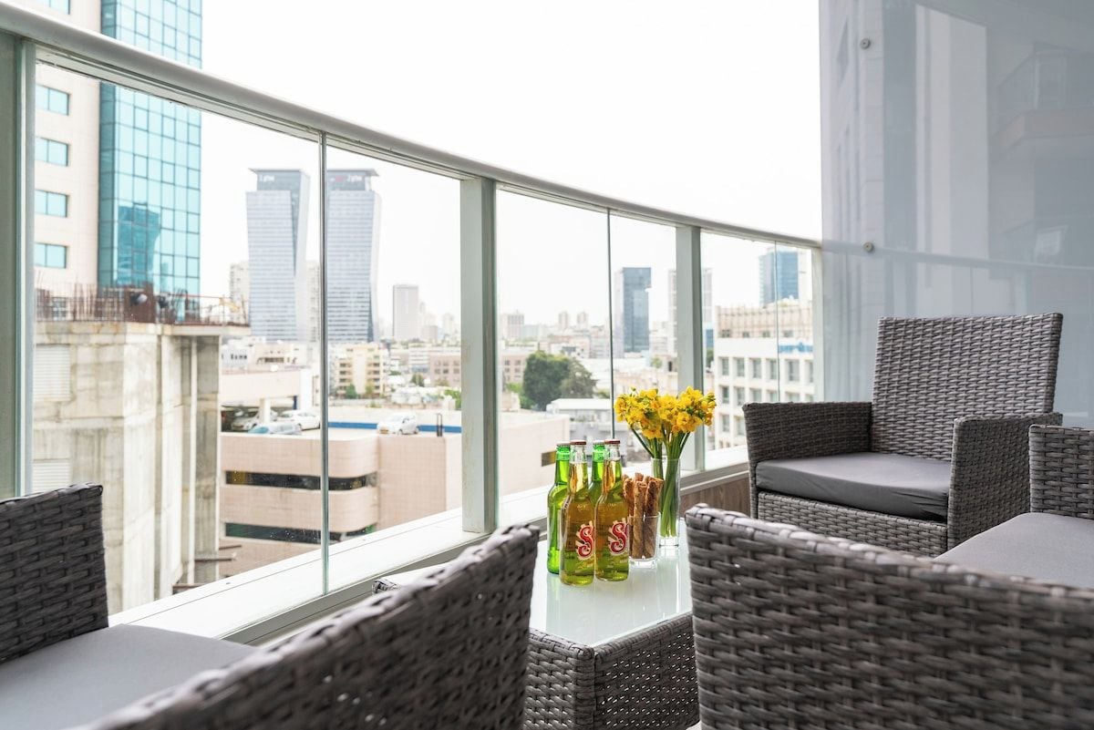 Un balcón con mobiliario de mimbre, una mesa con botellas y flores, y vista a la ciudad a través de amplias ventanas.
