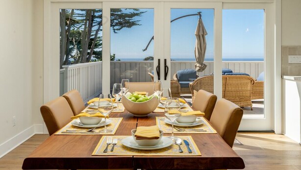Un coin repas avec une table en bois dressée pour quatre personnes, un saladier rempli de fruits et une vue sur l’océan par de grandes portes vitrées.