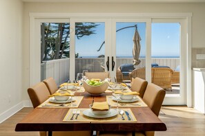 A dining area with a wooden table set for four, a bowl of fruit, and a view of the ocean through glass doors.