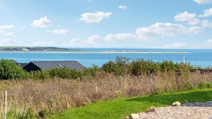 Een kustlandschap met een grasveld, een klein gebouw met een donker dak, en uitzicht op de zee en verafgelegen land.