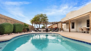 A swimming pool with a wooden pergola, a covered seating area, and a view of a building with a beige facade.