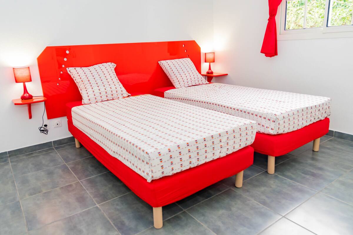 A room with two beds, each with a red headboard and white bedding with a pattern. There are two bedside tables with lamps, and a window with a red curtain.