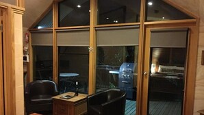 Een kamer met een grote schuifglazen deur die uitkomt op een terras. Binnen staat een houten salontafel, twee zwarte stoelen en een bijzettafeltje met een fles en een boek.