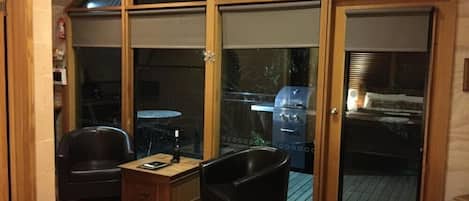 Una habitación con un amplio ventanal corredera que da a una terraza exterior. En el interior, hay una mesa de centro de madera, dos sillas negras y una mesita con una botella y un libro.