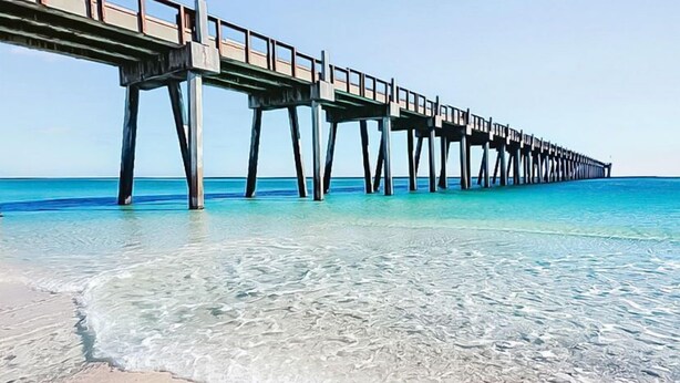 Un molo che si protende su un mare cristallino, con una spiaggia sabbiosa in primo piano.