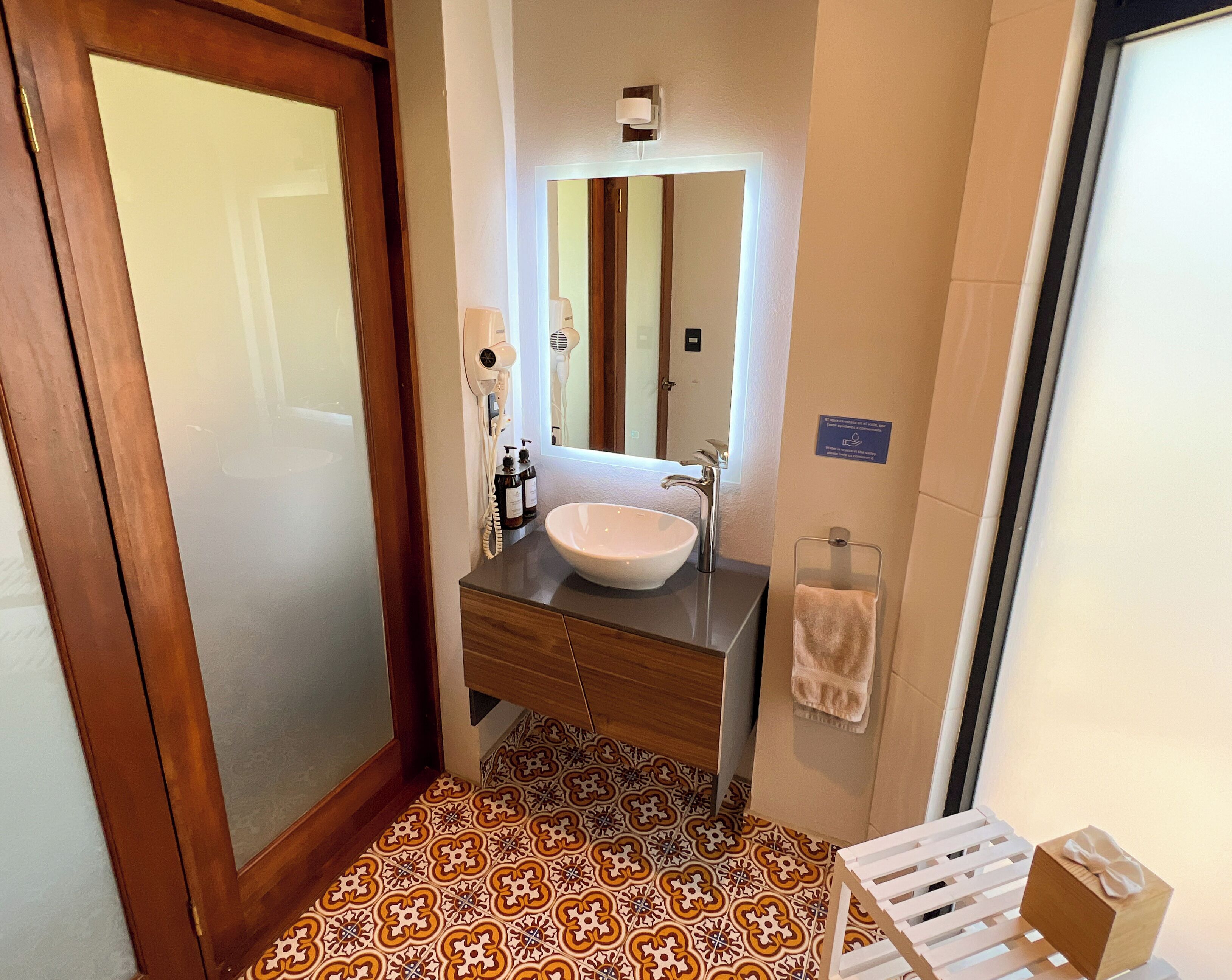 A bathroom with a wooden vanity, white sink, and a mirror. There is a towel rack with a towel and a small gift box on a white stool. The floor has patterned tiles.