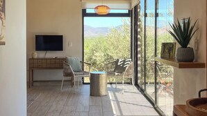 Una sala de estar moderna con una televisión de pantalla plana, un sofá y un sillón. Se aprecia un amplio ventanal con vistas a árboles y montañas.