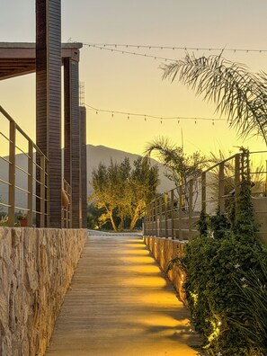 A pathway leading to a building with a stone wall on one side and a metal railing on the other, under a clear sky. - Valle Soléa (Valle de Guadalupe)