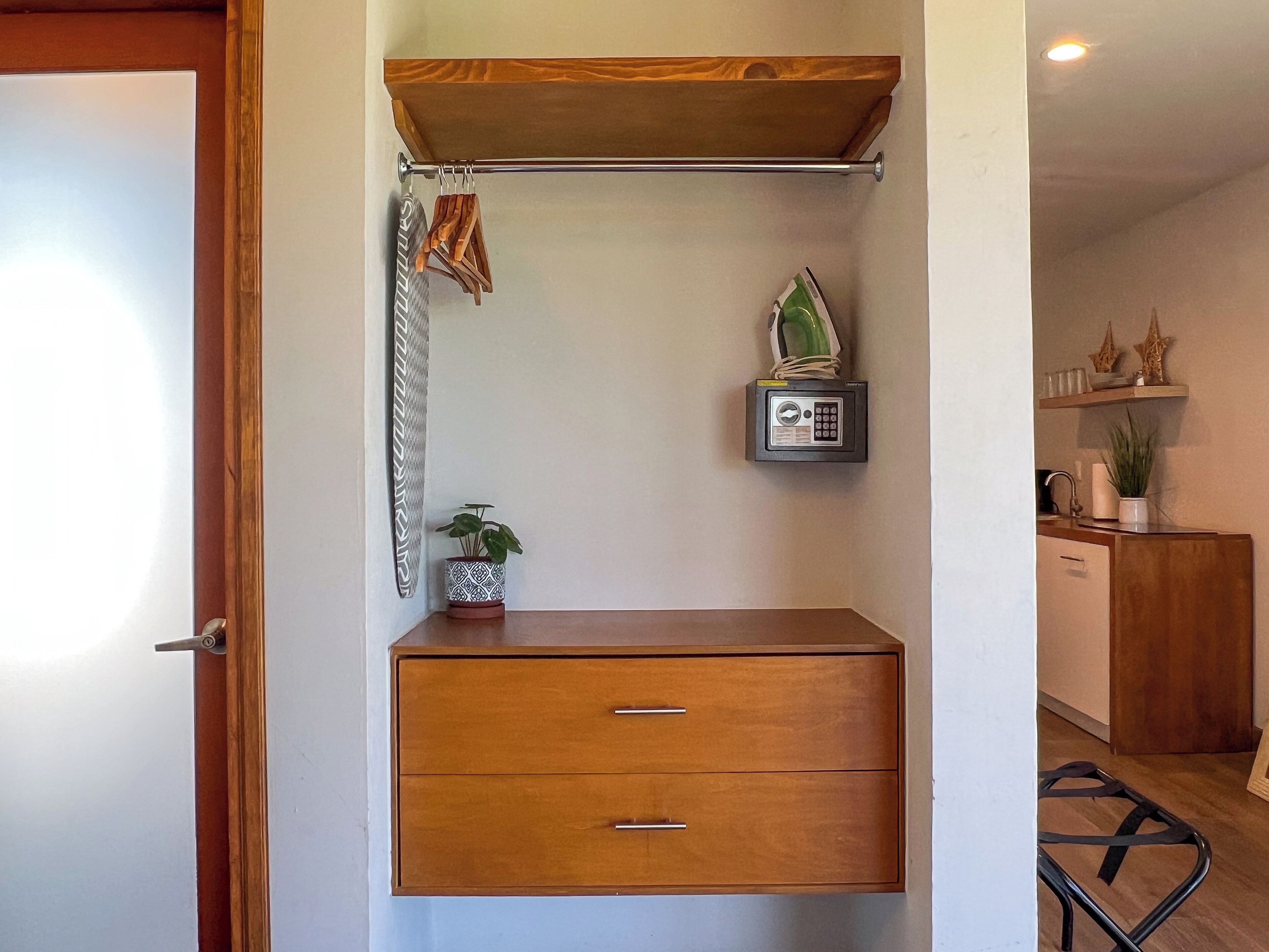 A small, neatly organized room with a wooden cabinet, a hanging coat rack, a potted plant, a small safe, and a shelf with decorative items.