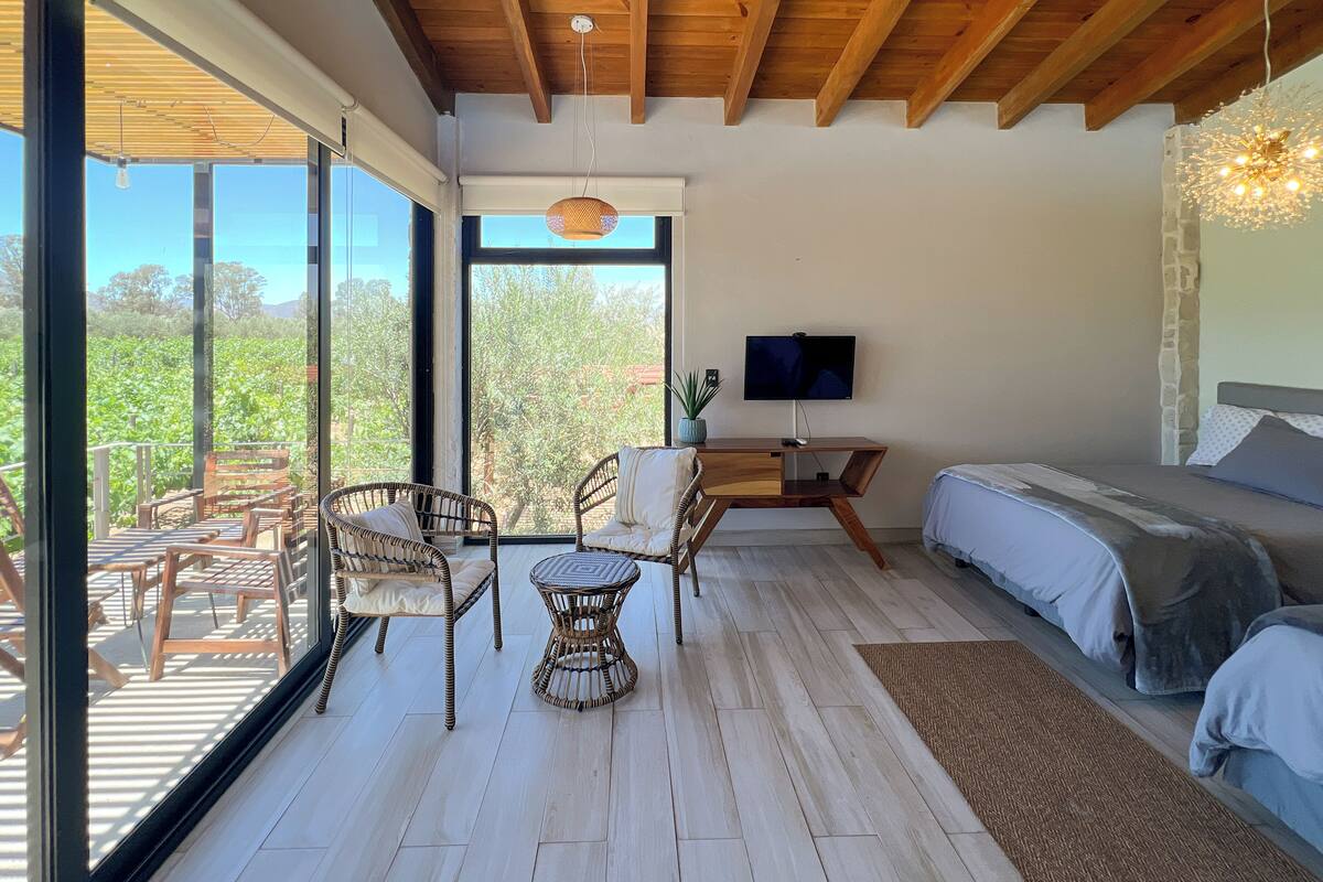 A bedroom with a bed, a TV, and a balcony with wicker furniture and a view of greenery.