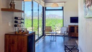 Amplia habitación con un gran ventanal corredero que da a un balcón con vistas a la montaña. Cuenta con un mueble de televisión de madera, una silla de mimbre y una pequeña mesa con estructura metálica.