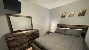 A bedroom with a wooden dresser, a bed with grey bedding, a nightstand with a lamp, and three wall art pieces. - O2 Suites Houma (Houma)