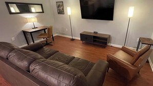 A living room with a brown leather sofa, a small wooden table with a lamp, a TV on the wall, and a floor lamp. - O2 Suites Houma (Houma)