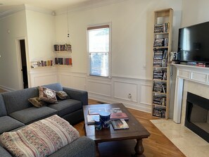 Un salon avec un canapé gris, une table basse sur laquelle sont posés des livres et une tasse, une cheminée et une bibliothèque remplie de DVD.