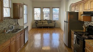 A kitchen with wooden cabinets, a stainless steel refrigerator, a stove, and a sink. There is a window with blinds and a sofa in the center of the room.