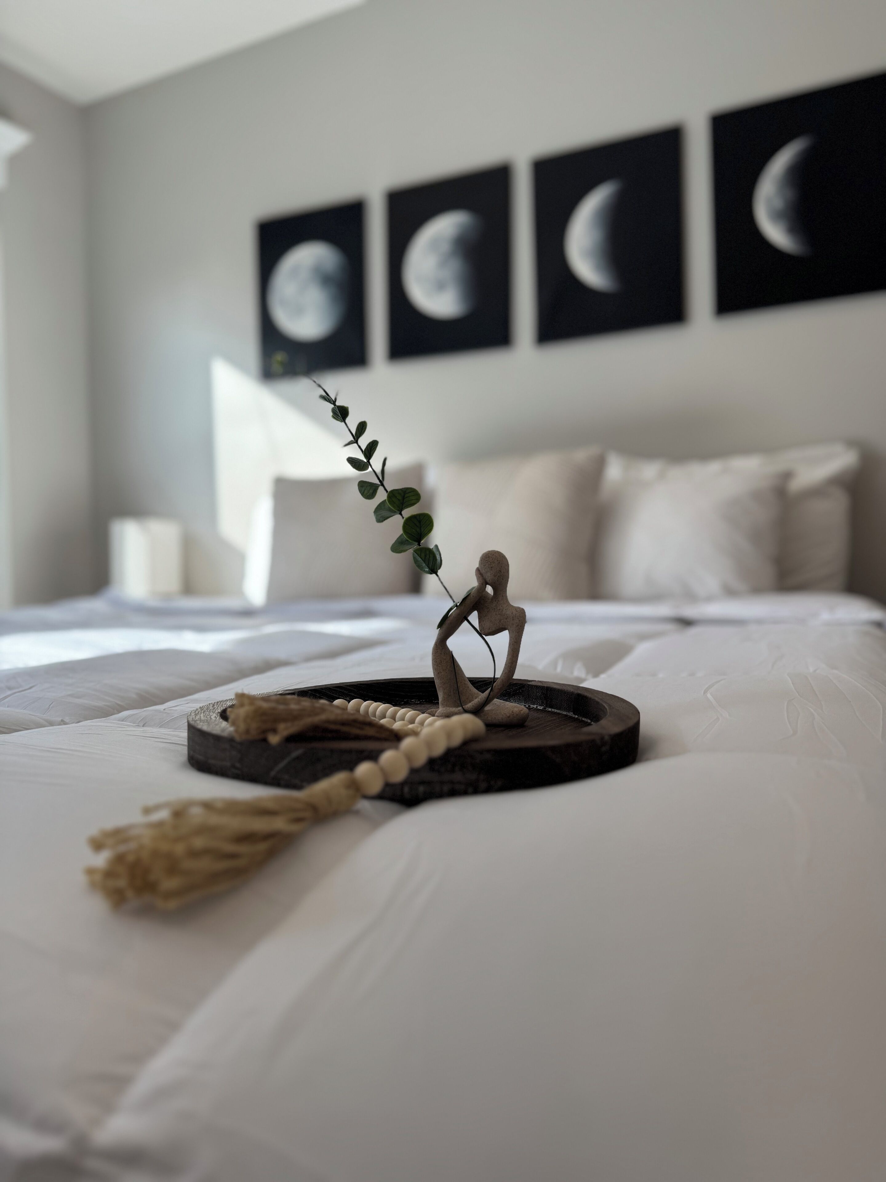 A neatly made bed with a decorative wooden figure, a beaded string, and a branch. There are four framed pictures of the moon on the wall.