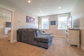 A living room with a grey sectional sofa, a small table, and a cabinet. There are two windows and a world map on the wall. - Downtown Extended Stay ~ Try Purple Mattress Brand - Middlesex County (South Plainfield)