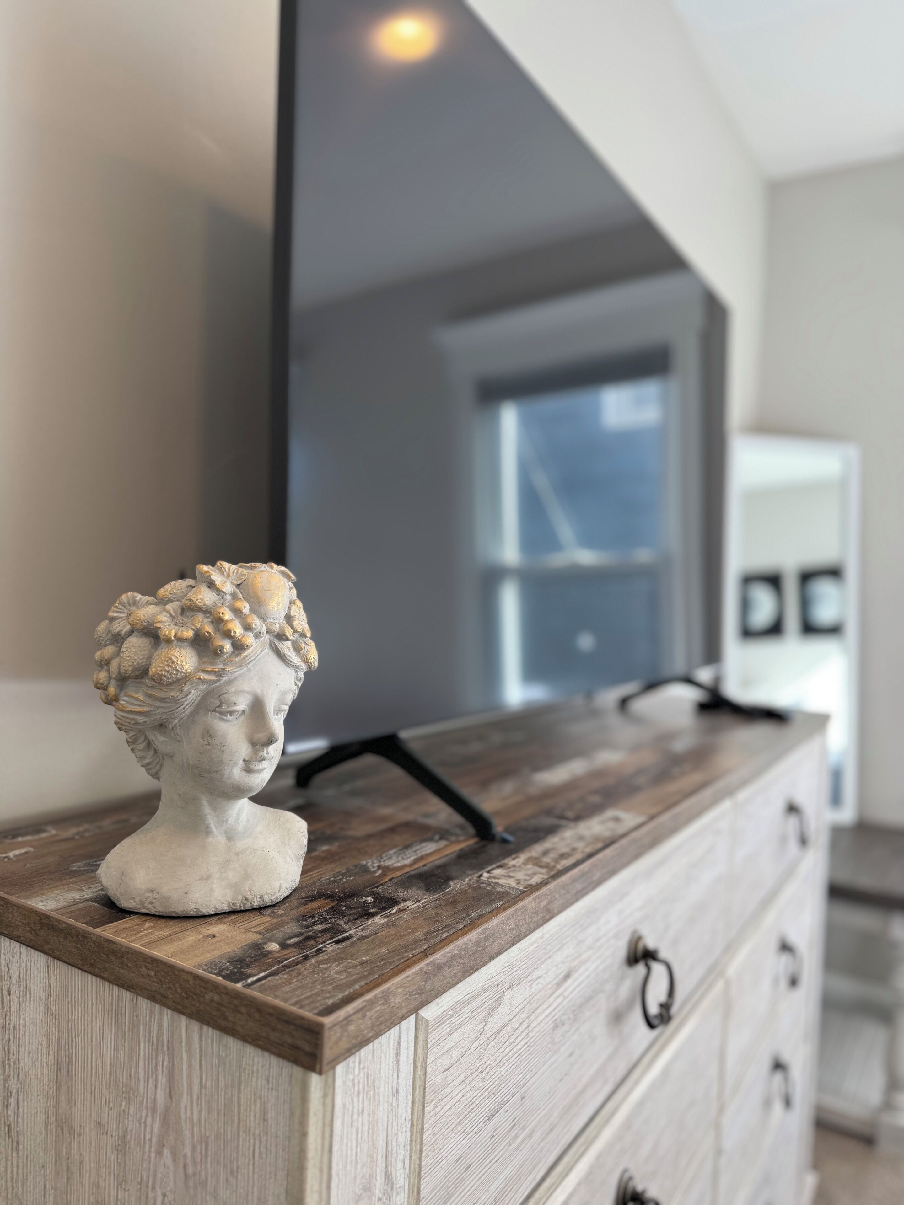 Un aparador de madera con un busto de mármol encima, un espejo arriba y una ventana y una puerta al fondo.