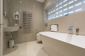 A modern bathroom with a large bathtub, a sink, and a toilet. There are shelves with toiletries and a towel rack.