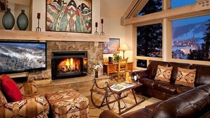 Una sala de estar acogedora con chimenea, asientos cómodos, televisión y un ventanal con vistas a un pueblo nevado.