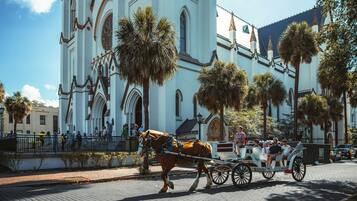 Une calèche tirée par des chevaux sur une rue pavée, avec en arrière-plan un bâtiment blanc doté d’arches et d’un clocher.