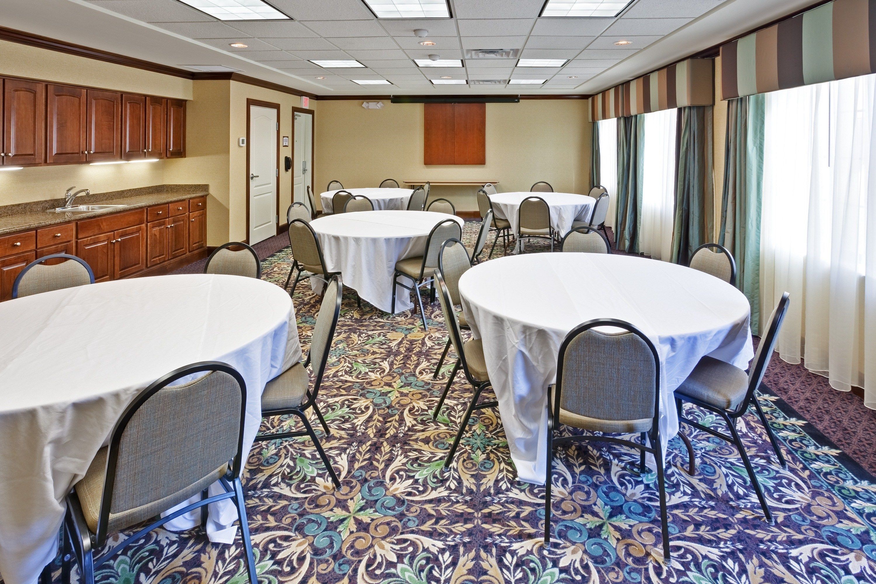 A conference room with round tables and chairs, a kitchenette, and a patterned carpet.