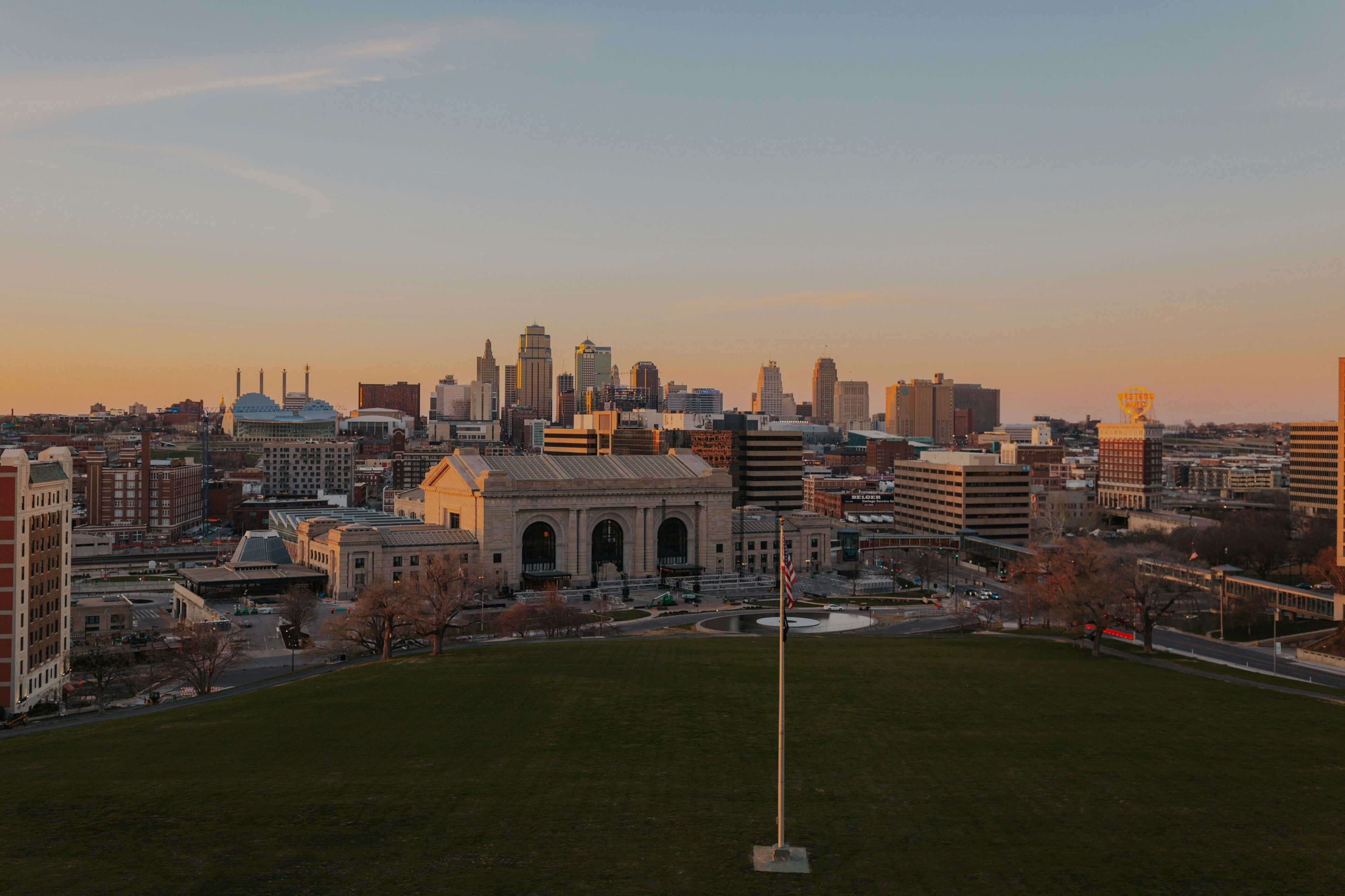 A city skyline with a prominent building with a dome and arched entrance, surrounded by various other buildings and a grassy area in the foreground.