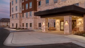 A multi-story hotel with a modern design, featuring a covered entrance with benches and exterior lighting. - Flexible 1-Bedroom Suite w/ Full Kitchen, KS (Overland Park)