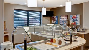 A buffet area with a glass partition, various food items, condiments, and a television mounted on the wall. - Flexible 1-Bedroom Suite w/ Full Kitchen, KS (Overland Park)