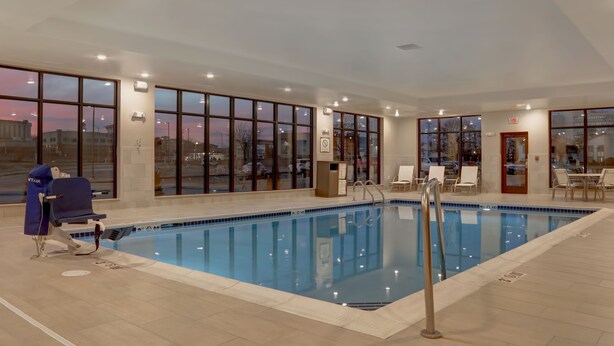 A swimming pool with clear blue water, surrounded by large windows, and a chair with a towel. - Flexible 1-Bedroom Suite w/ Full Kitchen, KS (Overland Park)
