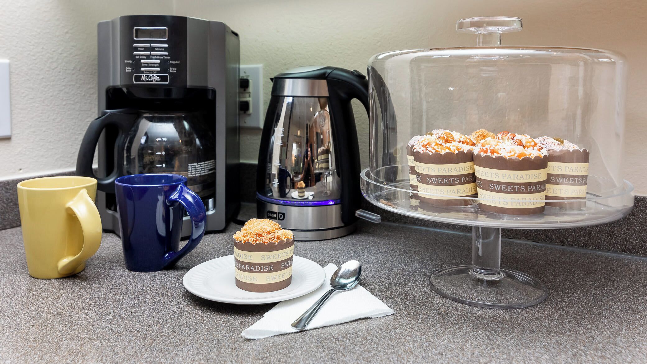 Eine Küchenarbeitsplatte mit Kaffeemaschine, Wasserkocher, einer Kuchenplatte mit Desserts und zwei Tassen.